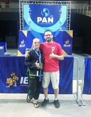 Woman in black gi with a medal and a bearded man in red shirt pose in front of a PAN IBJJF championship backdrop