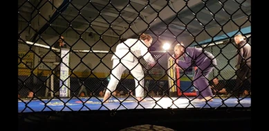 Two grapplers in white and purple gis face off on blue mats inside a caged enclosure during a competition