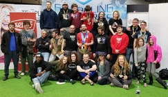 Large group of teens and adults, several wearing medals, pose together on green turf at a grappling tournament