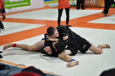 Two competitors in dark no-gi attire grapple on orange and white tournament mats, one holding the other down from the top