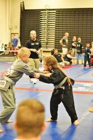 Two kids in gray and black gis grip each other while competing on blue mats as a referee watches in a gym