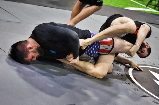 Two men grappling without gis on gray mats, one in a black rash guard, the other in stars-and-stripes shorts