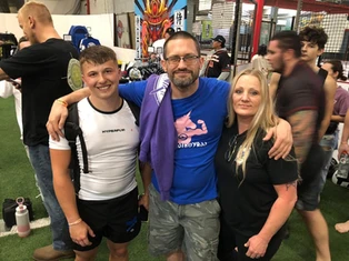 A smiling young man, a bearded man in a blue shirt and a blonde woman pose arm in arm at an indoor event