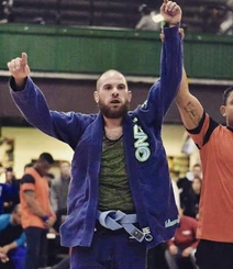 Bearded competitor in a blue gi has his arm raised by a referee in an orange shirt at a tournament