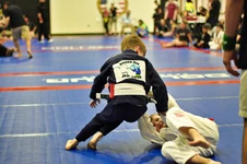 Two young children in gis grappling on blue tournament mats, one in a dark gi leaning over a child in white