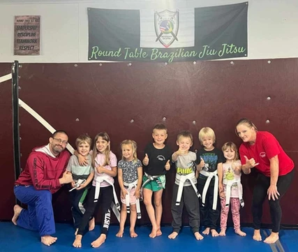 Two adults and a row of young kids give thumbs up under a Round Table Brazilian Jiu Jitsu banner