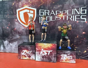 Three young boys wearing medals stand on a 1st, 2nd and 3rd place podium in front of a Grappling Industries backdrop
