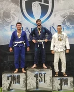 Three adult grapplers in gis wearing medals stand on a 1st, 2nd and 3rd place podium at a tournament