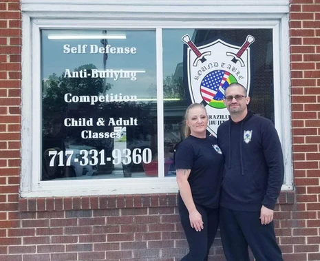 A woman and a man in black outside a brick building whose window lists self defense, anti-bullying and classes