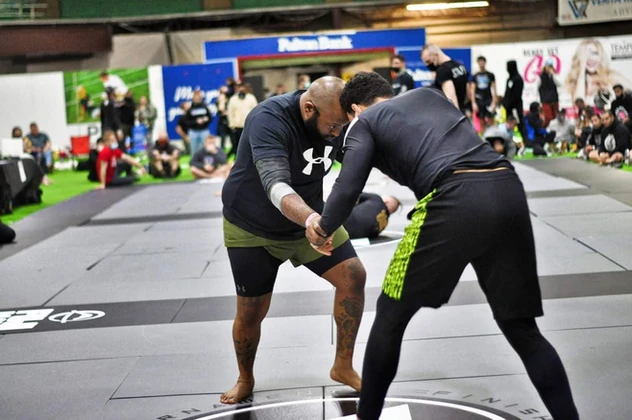 Two men gripping hands while standing on gray mats at a tournament, with spectators watching from the sidelines