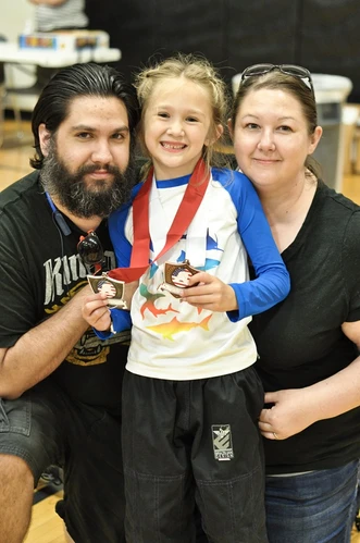 Smiling young girl in a shark-print rash guard holding two medals, posing between a bearded man and a woman
