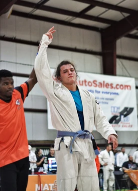 Referee in an orange shirt raising the arm of a competitor in a white gi at an indoor tournament