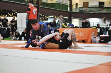 No-gi competitor pinning an opponent on the mat while a referee in orange watches at an indoor tournament