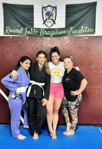 Four women in gis and casual clothes posing on blue mats under a Round Table Brazilian Jiu Jitsu banner