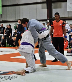Two gi competitors in a standing clinch and takedown attempt while a referee in orange looks on