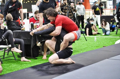 Competitor in a red shirt riding the back of a kneeling opponent on a mat in an indoor turf venue