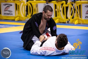 Competitor in a black gi kneels over an opponent in a white gi during a match on blue mats at an IBJJF event