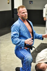 Bearded man in a blue gi and black belt kneels on the mat, gesturing as he explains a technique