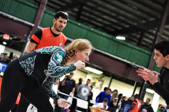 Woman with a long braid in a green and black rash guard squares up to an opponent at a tournament, referee behind