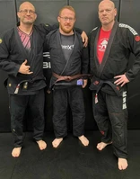 Three men in dark gis stand together smiling on dark gym mats, with their arms around one another