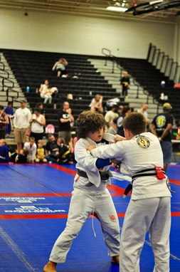 Two kids in white gis grip each other on blue mats at a tournament, spectators in the bleachers behind