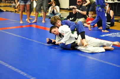 Young competitor in a blue gi controls an opponent in a white gi on the mat as spectators watch