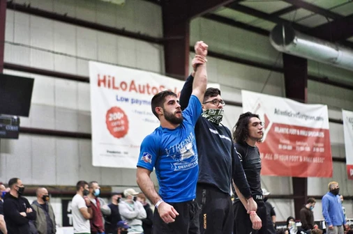 Referee raises the arm of a winner in a blue shirt after a no-gi match, sponsor banners behind