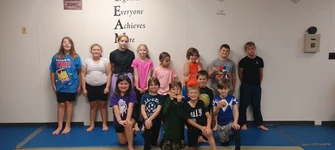 Group of kids in casual clothes pose on blue mats in front of a wall reading Everyone Achieves More