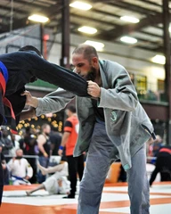 Bearded competitor in a gray gi grips the sleeve of an opponent in a dark gi on a tournament mat