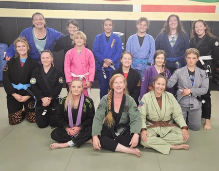 Group of women in colorful gis and belts smile together on the mats, some standing and some seated