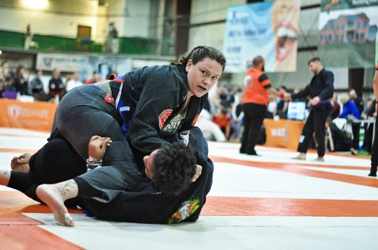 Competitor in a black gi controls an opponent on the mat during a jiu-jitsu tournament match with spectators behind