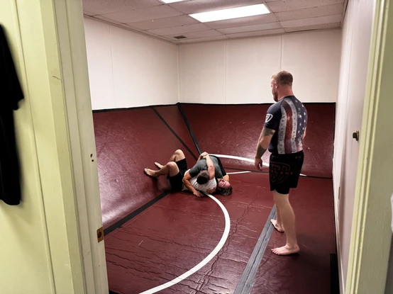 Two people grappling on the floor of a maroon-matted room while an instructor in an American flag rash guard watches