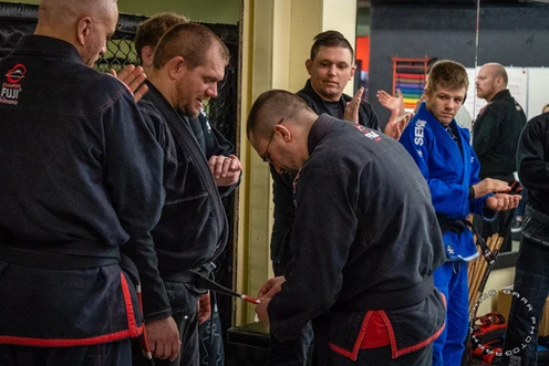 Group of men in dark gis and one in a blue gi gather around as an instructor handles a student's belt