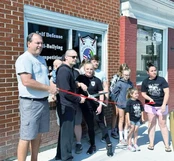 Adults and children gather outside a brick storefront as a woman cuts a red ribbon with large scissors