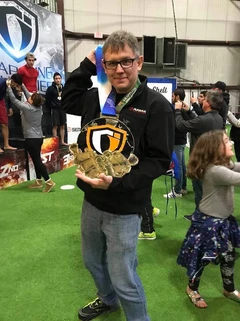 Man with glasses holds up a large gold tournament medal on a green indoor turf floor with onlookers behind him