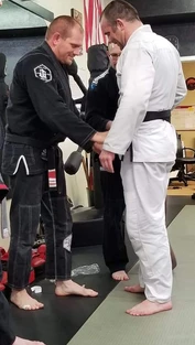 Instructor in a black gi adjusts the black belt of a man in a white gi while another person stands behind