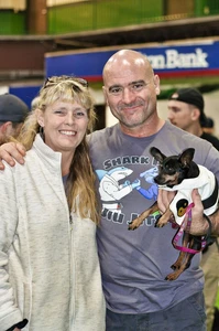 Smiling woman and bald man in a Shark Pit Jiu Jitsu shirt posing with a small dog in a white jacket