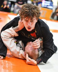 Curly-haired competitor in a black gi pins an opponent in a white gi on orange mats during a tournament match
