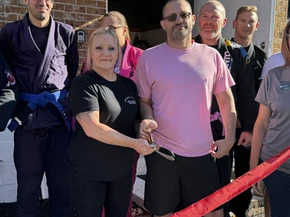 Woman in black shirt and man in pink shirt cut a red ribbon with scissors as gi-wearing students look on