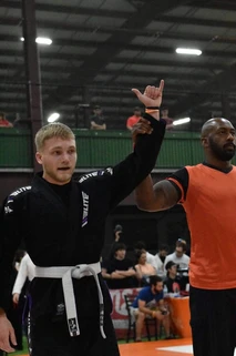 Referee in an orange shirt raises the arm of a competitor in a black gi and white belt at an indoor tournament