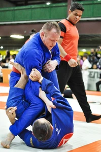 Two competitors in blue gis grapple on tournament mats, one in open guard, as a referee in orange watches