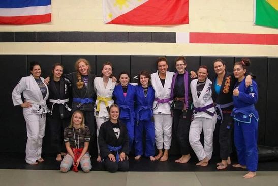Group of women in gis of various colors and belts pose in front of a padded wall with flags