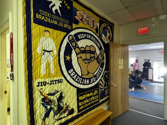 Large Brazilian Jiu-Jitsu quilt reading Keep on Rolling hangs in a hallway beside a doorway to the mat room