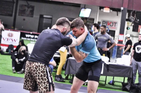 Two no-gi competitors grip up in a standing clinch on a turf-floored tournament area with spectators behind