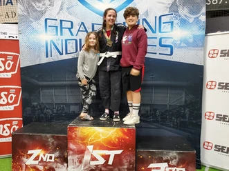Woman wearing a medal and gi stands on a first-place podium with two children before a Grappling Industries backdrop