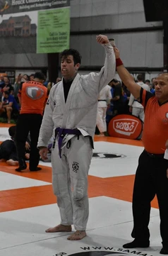 Referee raises the arm of a competitor in a white gi and purple belt on orange and white tournament mats