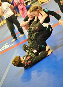 Two young competitors in dark gis grapple on blue mats, one on their back in guard, as a referee crouches nearby