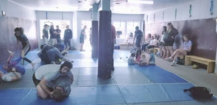 Several pairs grapple on blue mats in a gym while spectators sit along the wall and benches