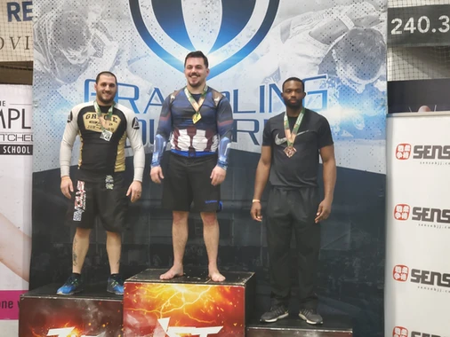 Three men wearing medals stand on a podium in front of a grappling tournament backdrop