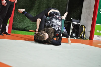 Two competitors in dark rash guards grappling on a mat during a tournament match, one on top of the other